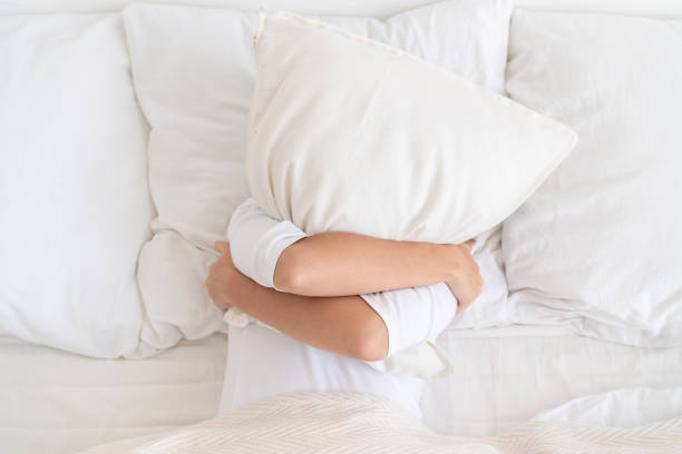 jovencita abrazando almohada, cubriéndose la cara mientras se acuesta en la cama con camiseta blanca, divirtiéndose, no listo para levantarse, relajándose tarde en la mañana - late afternoon fotografías e imágenes de stock