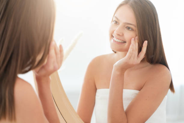 młoda kobieta owinięta w ręcznik kąpielowy stojący przed lustrem w łazience, dotykając jej skóry twarzy, masując, uśmiechając się szczęśliwie - clear sky human skin towel spa treatment zdjęcia i obrazy z banku zdjęć