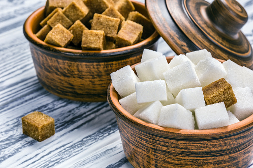 White refined sugar and brown unrefined sugar cubes in ceramic bowls on a light wooden table.