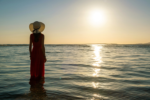 Young woman wearing long red dress and straw hat standing in sea water at the beach enjoying view of rising sun in early summer morning.