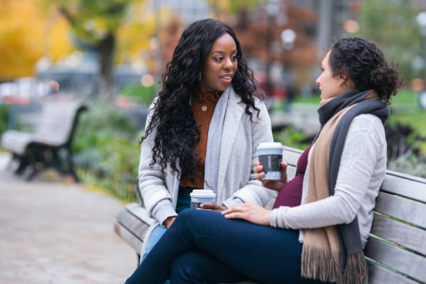 беременная женщина разговаривает с подругой во время отдыха на скамейке в парке - human pregnancy african ethnicity women family стоковые фото и изображения
