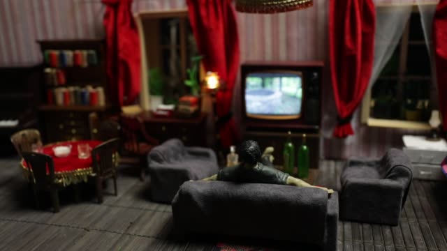 A realistic dollhouse living room with furniture and window at night. Artwork table decoration with handmade realistic dollhouse. Man wathing retro style Television in dark room. Selective focus.