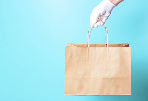 la mano femenina en un guante blanco sostiene bolsa de cartón marrón sobre un fondo azul, concepto de entrega de alimentos. - box blank brown white fotografías e imágenes de stock