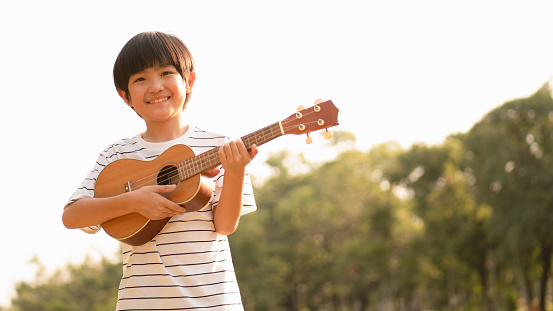 Cute Asian boy holding Ukulele with a big smile feeling happy and confident to play music in the park, carefree mind, concept musical playing, creative activity, music education, children development.