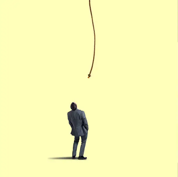 A businessman places his hands in his pockets as he leans back and looks up at a rope that dangles down from above.