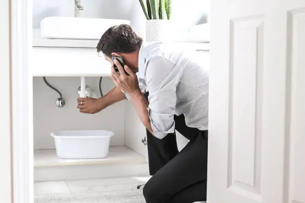 Photo of Sad Young Man Calling Plumber In Front Of Water Leaking From Sink Pipe