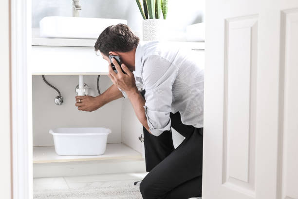 sad young man calling plumber in front of water leaking from sink pipe - home damage imagens e fotografias de stock