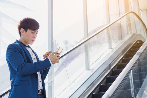 istock Asian handsome young business smiling and holding smartphone while standing on the escalator. Business and finance concept. 1218689685