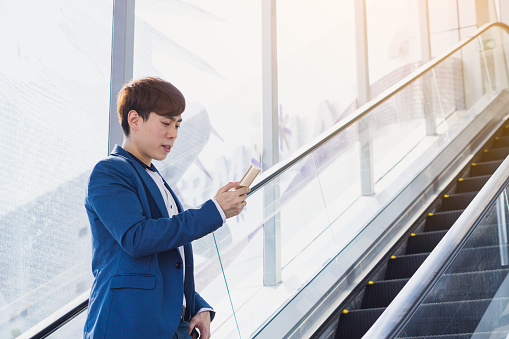 Asian handsome young business smiling and holding smartphone while standing on the escalator. Business and finance concept.