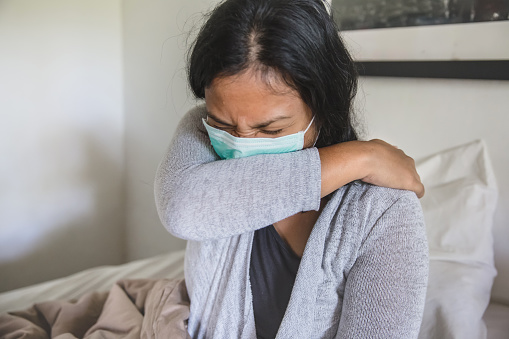 Portrait shot of Asian woman sick in bed,  covering cough with elbow, during home quarantine Covid 19