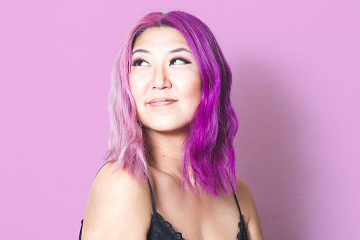 A Korean woman with pink and purple hair against a pink background looks off-camera with a slight smile