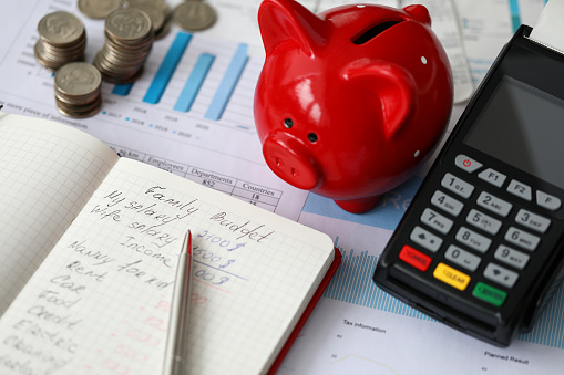 Close-up of red piggy bank terminal and notebook with notations. Monthly expenses on rent car food and credit. Cash money on desktop. Family budget concept
