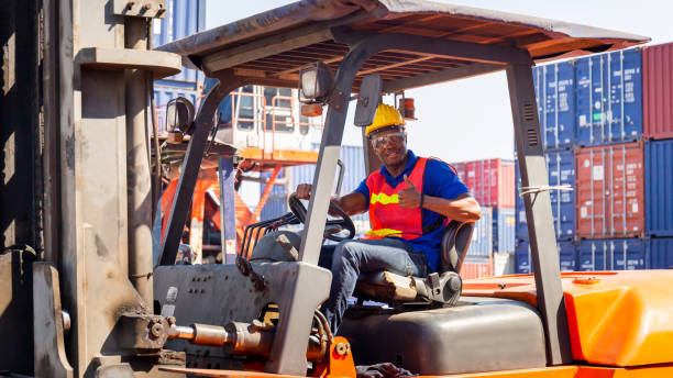 istock Worker man in hardhat and safety vest sitting in container stackers smiling with giving thumbs up as sign of Success 1218702055
