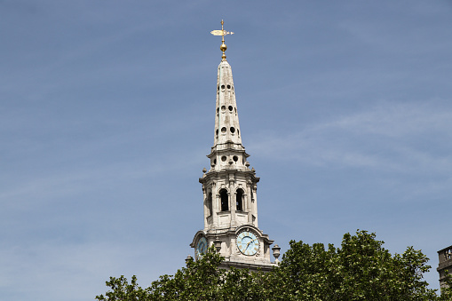 Forgotten church steeple between trees in London city. Historical monument from middle ages. Tower ends with a beautiful golden bird. In the middle there is wondeful clock with blue background.