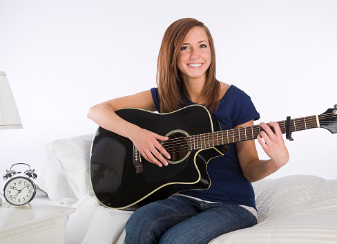 A young woman sitting on a bed playing guitar.