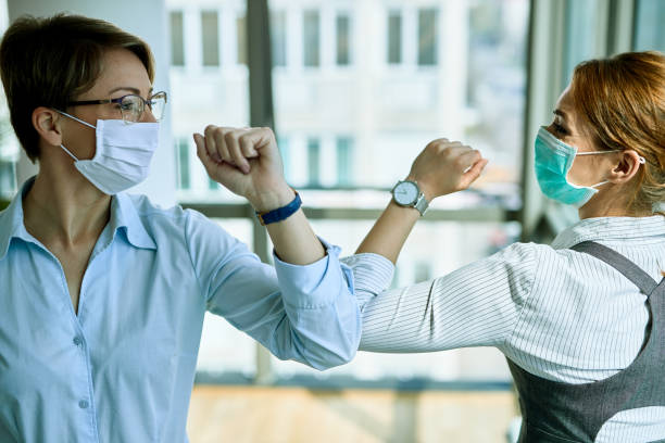 Business colleagues with face masks greeting with elbows during coronavirus pandemic. Two businesswomen elbow bumping while greeting each other in the office during COVID-19 epidemic. avoidance photos stock pictures, royalty-free photos & images