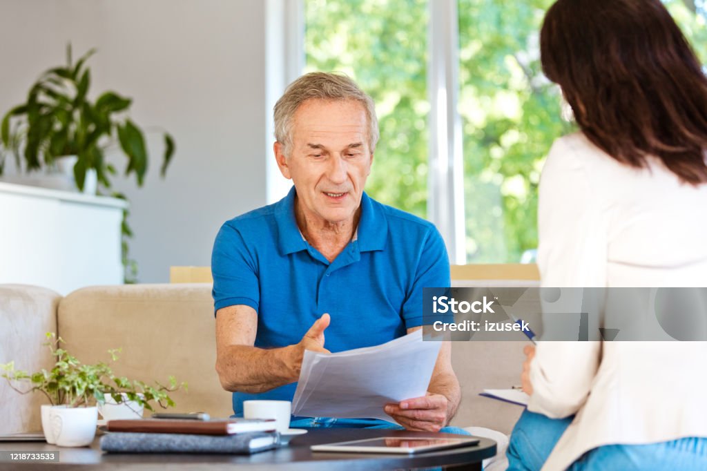 Displeased senior man having meeting with financial advisor at home Worried mature man sitting on sofa in living room at home. Senior man talking with female insurance agent. Senior Adult Stock Photo