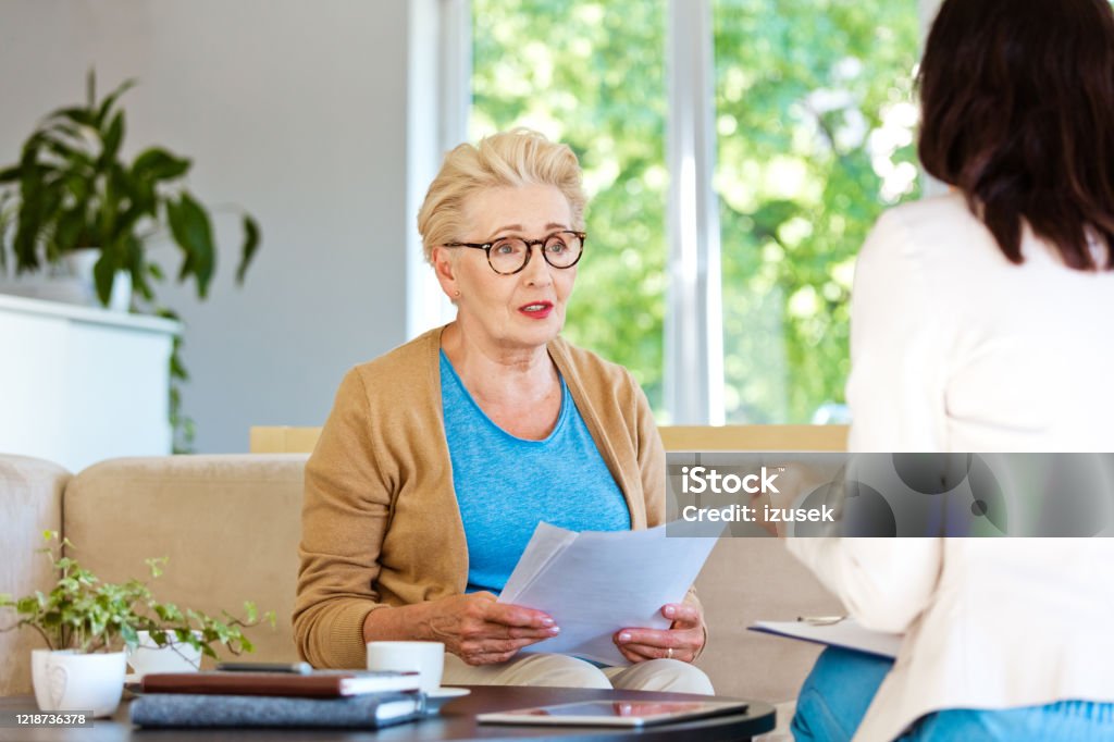 Worried senior woman having meeting with financial advisor at home Sad mature woman sitting on sofa in living room at home. Senior woman talking with female insurance agent. Asking Stock Photo
