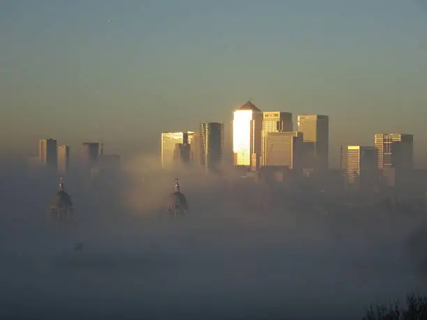 Fog sinks to reveal the skysrapers of Canary Wharf behind the Royal Navy Collage and Queens House.