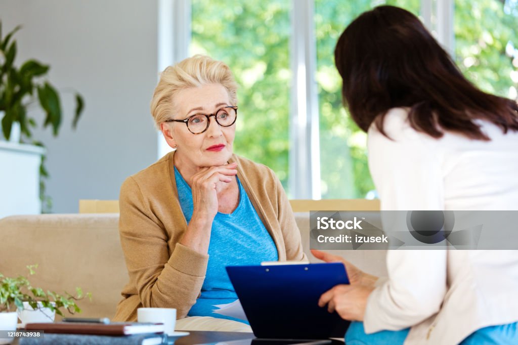 Insurance agent explaining agreement to senior woman Mature woman sitting on sofa in living room at home and listening to financial advisor. Insurance agent explaining document to elderly lady. Real Estate Agent Stock Photo