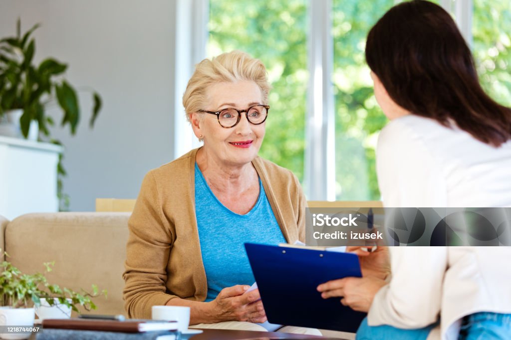 Insurance agent explaining agreement to smiling elderly lady Mature woman sitting on sofa in living room at home and listening to financial advisor. Insurance agent explaining document to senior woman. Active Seniors Stock Photo