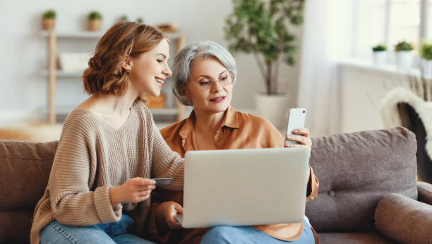 Young woman with mother shopping online at home Positive young female with gray haired mother sitting on sofa with laptop and using smartphone while performing online transactions with credit card during shopping old-phone stock pictures, royalty-free photos & images