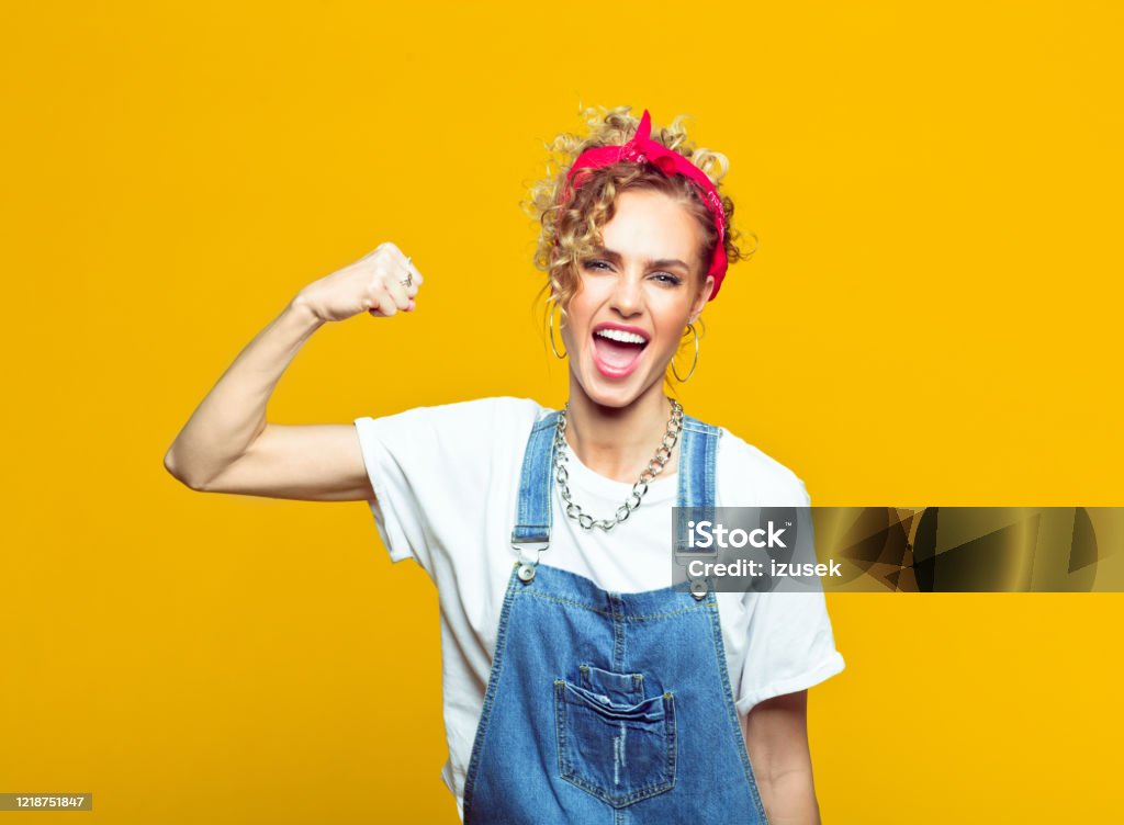 Powerful young woman in dungarees raising fist, portrait on yellow background Portrait of confident young woman wearing white t-shirt, denim dungarees and red bandana raising her fist and shouting at camera. Studio shot on yellow background. Repairing Stock Photo