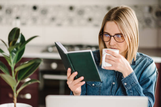 istock Home work. Girl drinking coffee and read a book at home. Woman working, learns, using laptop computer on table. Freelance. Paperwork. 1218758848