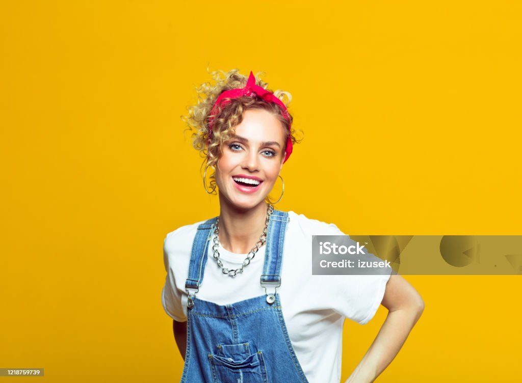 Cheerful young woman in 80's style outfit, portrait on yellow background Portrait of happy young woman wearing white t-shirt, denim dungarees and red bandana smiling at camera. Studio shot on yellow background. Women Stock Photo