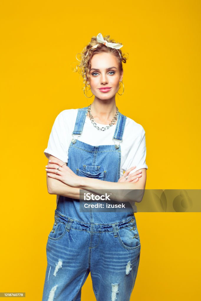 Portrait of confident young woman in coveralls on yellow background Portrait of young woman wearing white t-shirt, denim dungarees and bandana looking at camera with arms crossed. Studio shot on yellow background. 1980-1989 Stock Photo