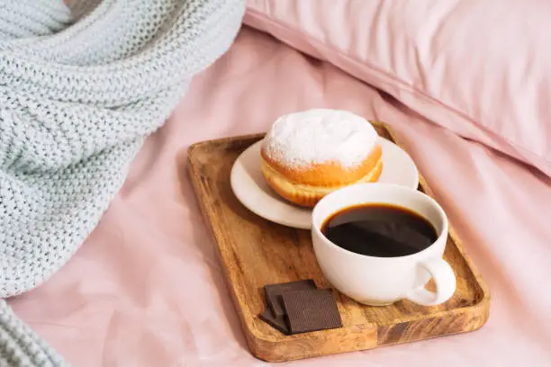 Morning breakfast in bed with fresh and hot coffee, a doughnut in powdered sugar and two slices of chocolate on a wooden tray against a backdrop of pink sheets and blue plaid. top view