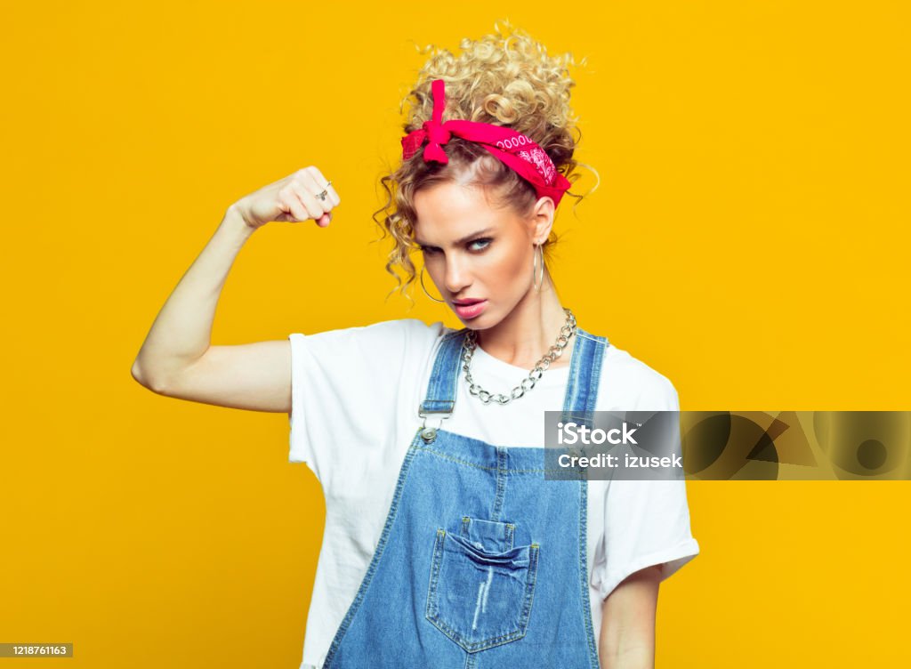 Powerful young woman in dungarees raising fist, portrait on yellow background Portrait of confident young woman wearing white t-shirt, denim dungarees and red bandana raising her fist and shouting at camera. Studio shot on yellow background. Hair Bun Stock Photo