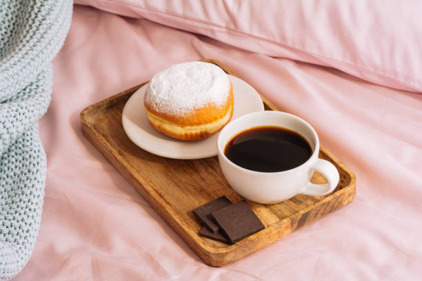 istock Morning breakfast in bed with fresh and hot coffee, a doughnut in powdered sugar and three slices of chocolate on a wooden tray against a backdrop of pink sheets and blue plaid 1218761199