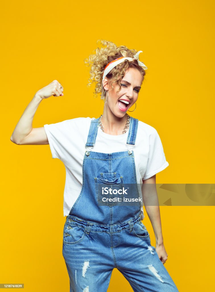 Powerful young woman in dungarees raising fist, portrait on yellow background Portrait of confident young woman wearing white t-shirt, denim dungarees and bandana raising her fist and shouting at camera. Studio shot on yellow background. DIY Stock Photo