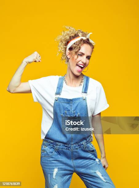 Powerful Young Woman In Dungarees Raising Fist Portrait On Yellow Background Stock Photo - Download Image Now