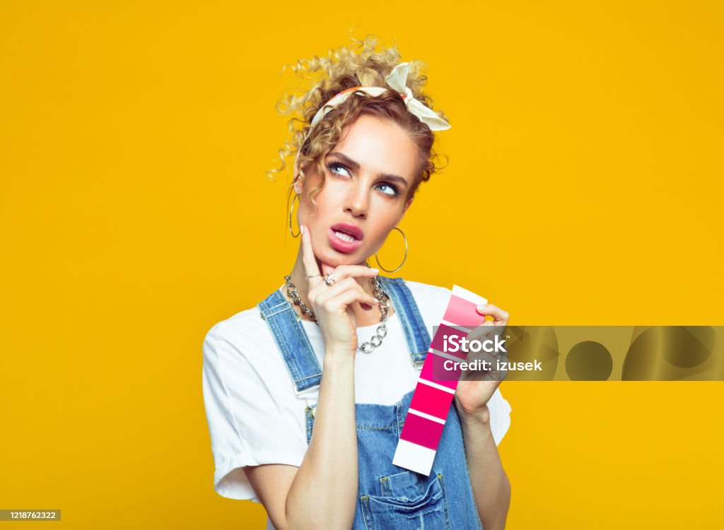 Young woman in coveralls holding color swatch Portrait of pensive young woman wearing white t-shirt, denim dungarees and bandana holding color swatch. Studio shot on yellow background. Choosing Stock Photo