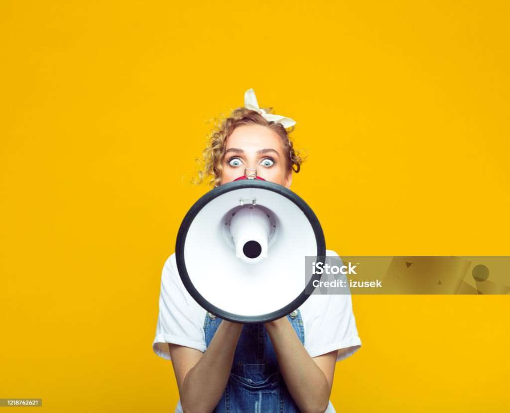 Young woman in dungarees shouting into megaphone Portrait of excited young woman wearing white t-shirt, denim dungarees and bandana shouting into megaphone. Studio shot on yellow background. Megaphone Stock Photo