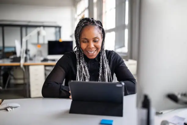 Smiling female executive sitting at her office desk making a video call with digital tablet. African american businesswoman working in office having a video conference.