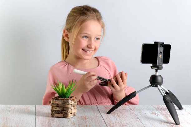 cute little blonde beauty blogger. girl speaking in front of the camera for vlog. a beautiful smiling schoolgirl sits at a table and talks about makeup - how to apply blush with a brush, on a little fashionista wearing a pink dress, on a gray background - child interview schoolgirl little girls imagens e fotografias de stock