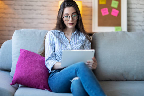 istock Pretty young woman using her digital tablet for reading while sitting on sofa at home. 1218766416