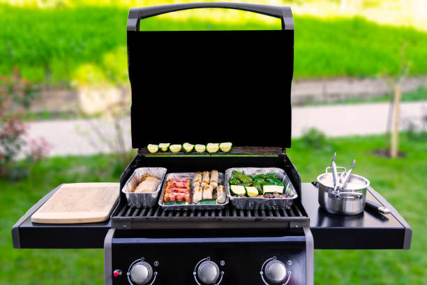 gas grill with vegetables and sausages An opened gas grill with vegetables, meat, and sausages in aluminum barbeque trays or dip pans. The background with green grass, bushes, a sidewalk are blurred. copy space on the black grill cover. barbecue grill stock pictures, royalty-free photos & images