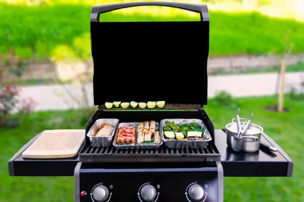 An opened gas grill with vegetables, meat, and sausages in aluminum barbeque trays or dip pans. The background with green grass, bushes, a sidewalk are blurred. copy space on the black grill cover.