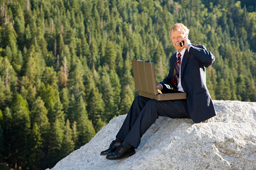 A businessman working on top of a rock in a wilderness area.