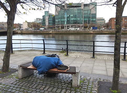 13th April 2020, Dublin, Ireland. Person sleeping on a River Liffey bench in Dublin city centre with the International Financial Services Centre IFSC in the distance across the river.