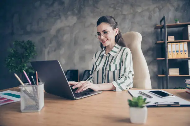 Portrait of positive cheerful freelancer lawyer girl sit desk work laptop, write online message start-up investors wear striped white shirt in workstation