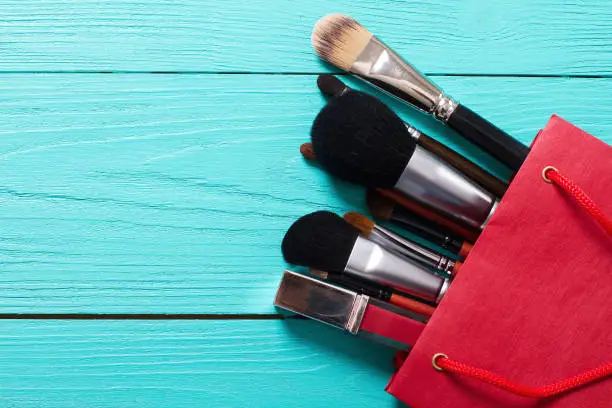 Photo of Makeup brushes on blue wooden background with copyspace. Make-up tools in red paper bag. Top view