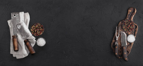 istock Vintage chopping board with meat hatchets and fork with knife on black background with salt and pepper. 1218796091