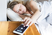 Young Eastern European woman lying in bed and turning off the alarm clock on her mobile phone in the morning