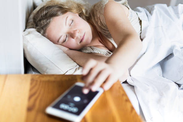 Young Eastern European woman lying in bed and turning off the alarm clock on her mobile phone in the morning stock photo