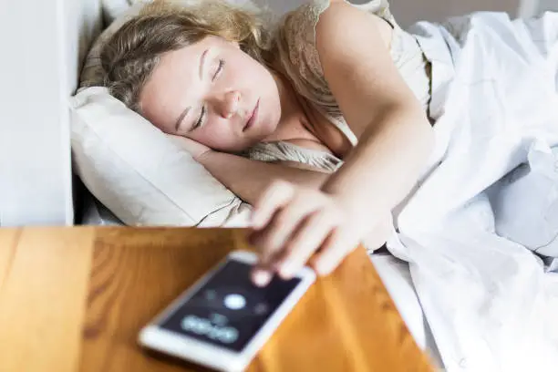 Young Eastern European woman lying in bed and turning off the alarm clock on her mobile phone in the morning.